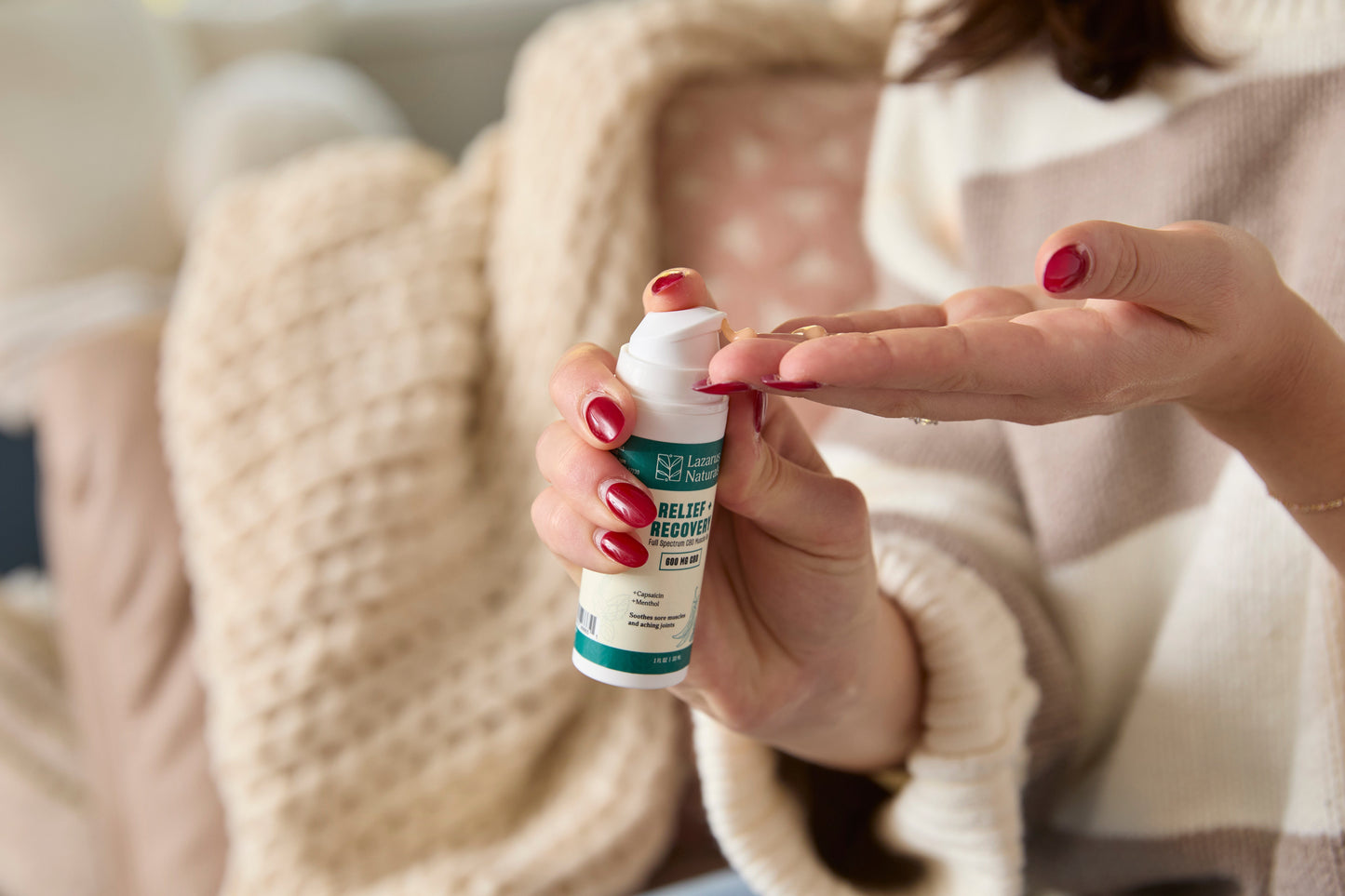 Woman sitting on couch putting Lazarus Naturals Relief + Recovery Muscle Gel into her hand
