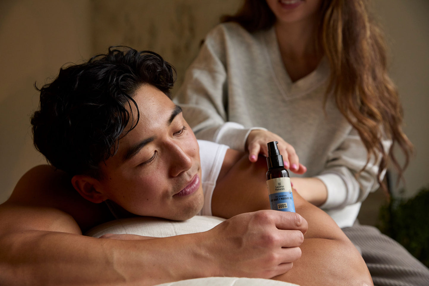 a man laying face down holding Lazarus Naturals massage oil while a woman is applying it to his shoulder.