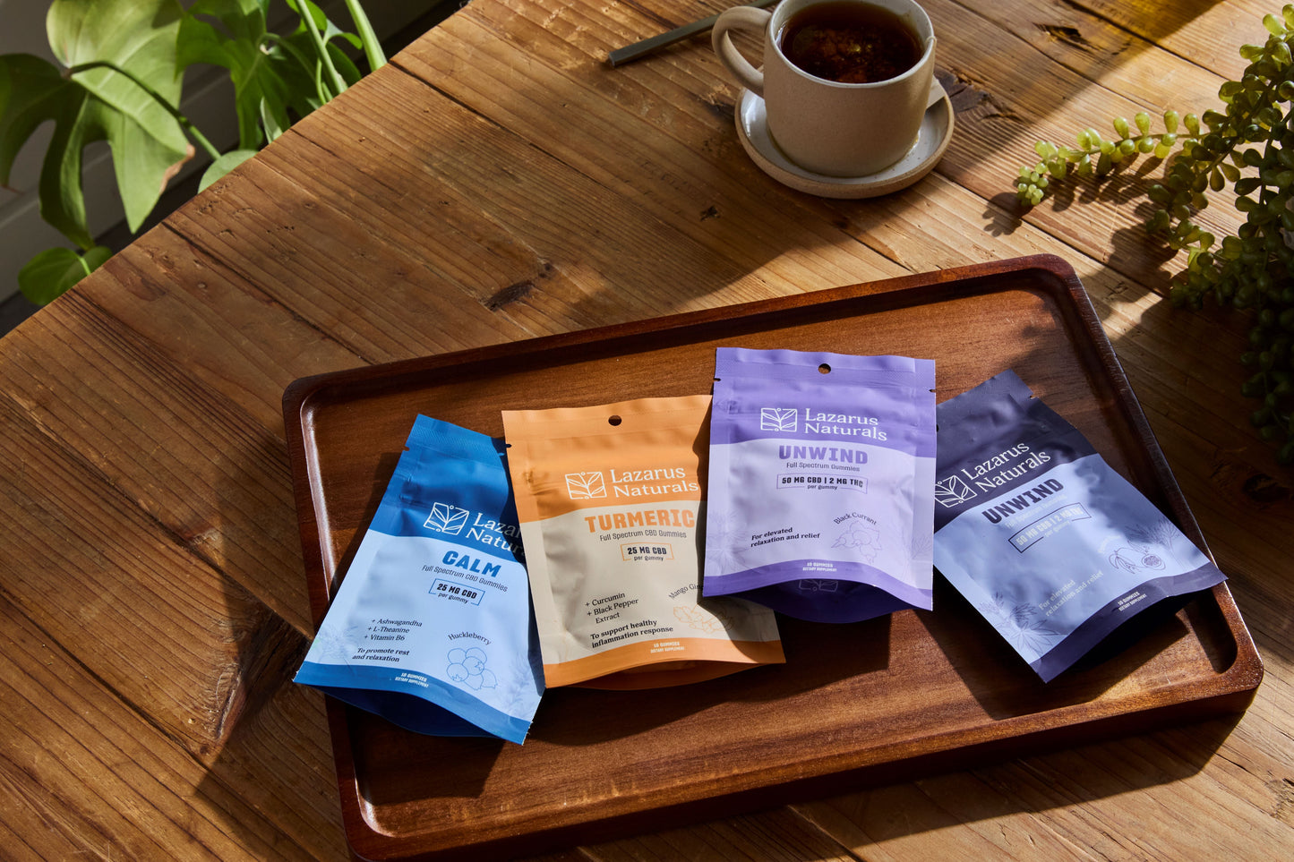 Lazarus Naturals Gummies in a row on a tray next to coffee. The gummies are: Calm Gummies, Turmeric, Unwind Black Currant Flavor, Unwind Passionfruit Flavor