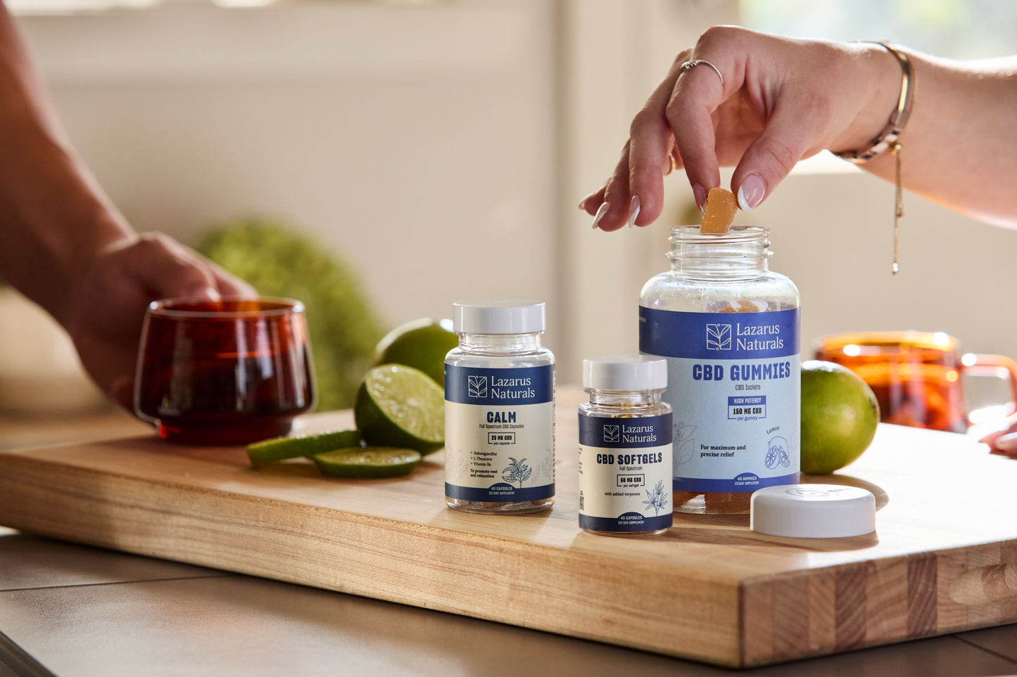 Person holding a cup on a cutting board with limes next to Lazarus Naturals Calm Capsules, Lazarus Naturals CBD Softgels, and a woman taking a Lazarus Naturals CBD Gummy out of the bottle. 